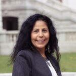 Smiling woman with dark hair and a blazer, photographed outdoors with stone steps in the blurred background.