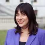 Portrait of a smiling woman with shoulder-length dark hair and bangs, wearing a purple blazer outdoors.